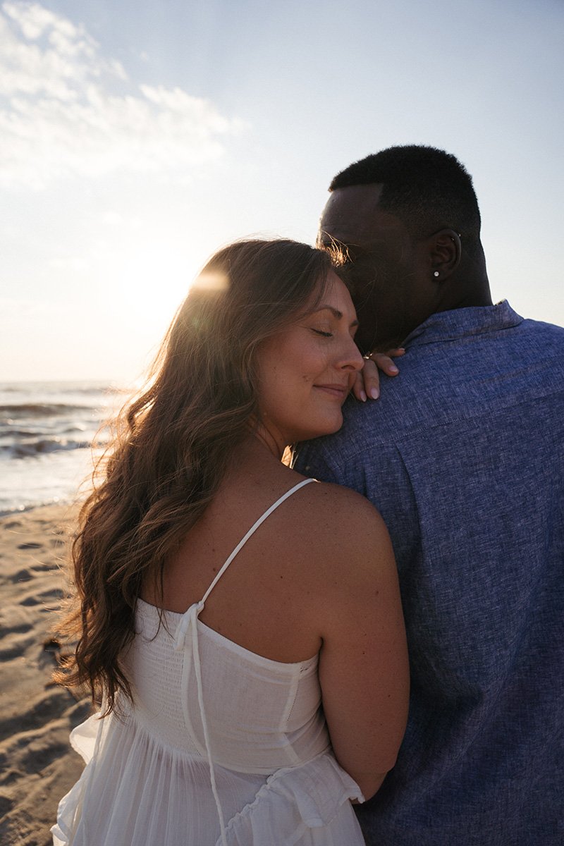 A couple embracing on the beach at sunset, with the woman smiling and the man holding her close.