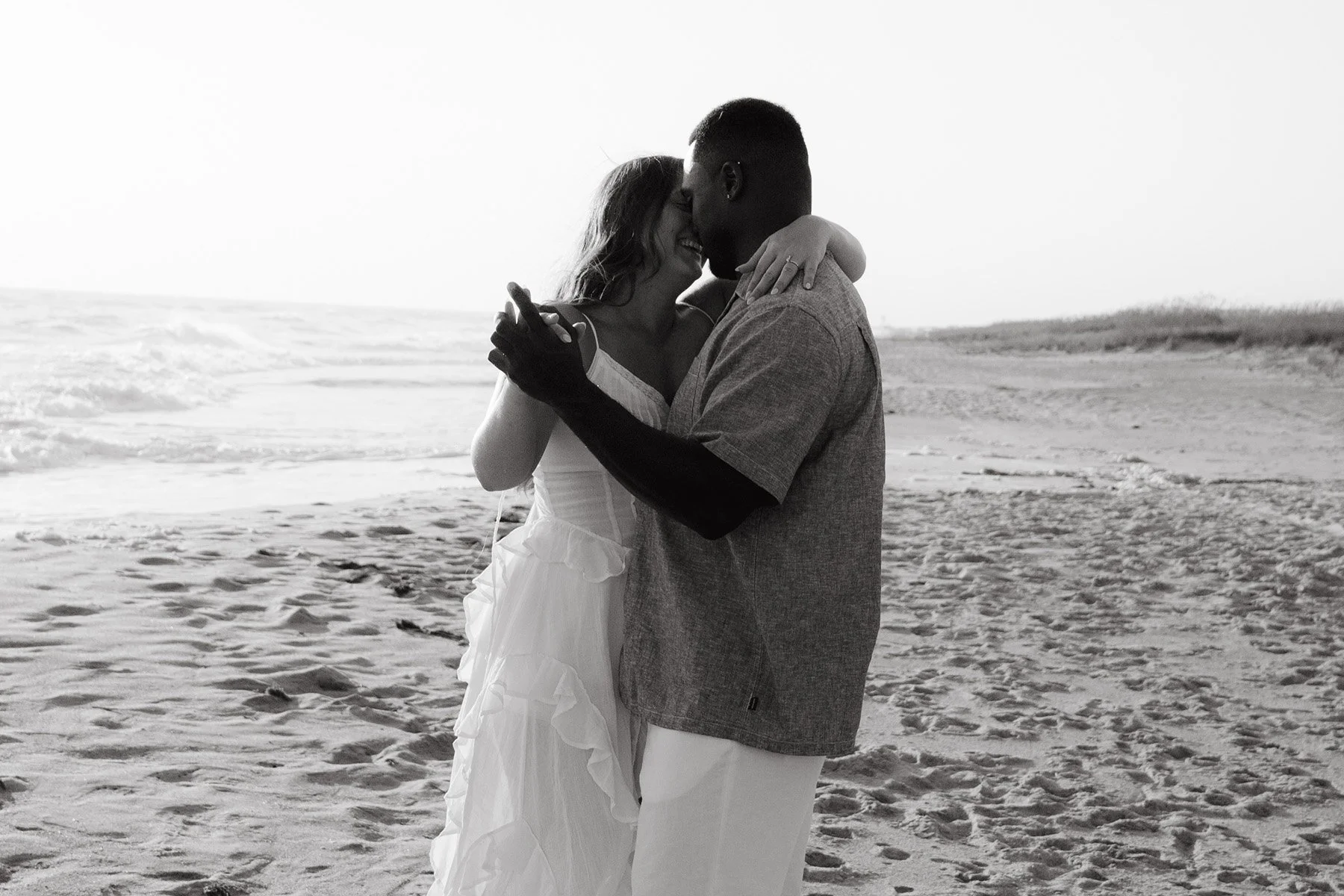A black and white photograph of a couple embracing on the beach, with the ocean behind them. The woman is wearing a white dress and the man is in a light shirt and white pants. They are smiling and touching noses.