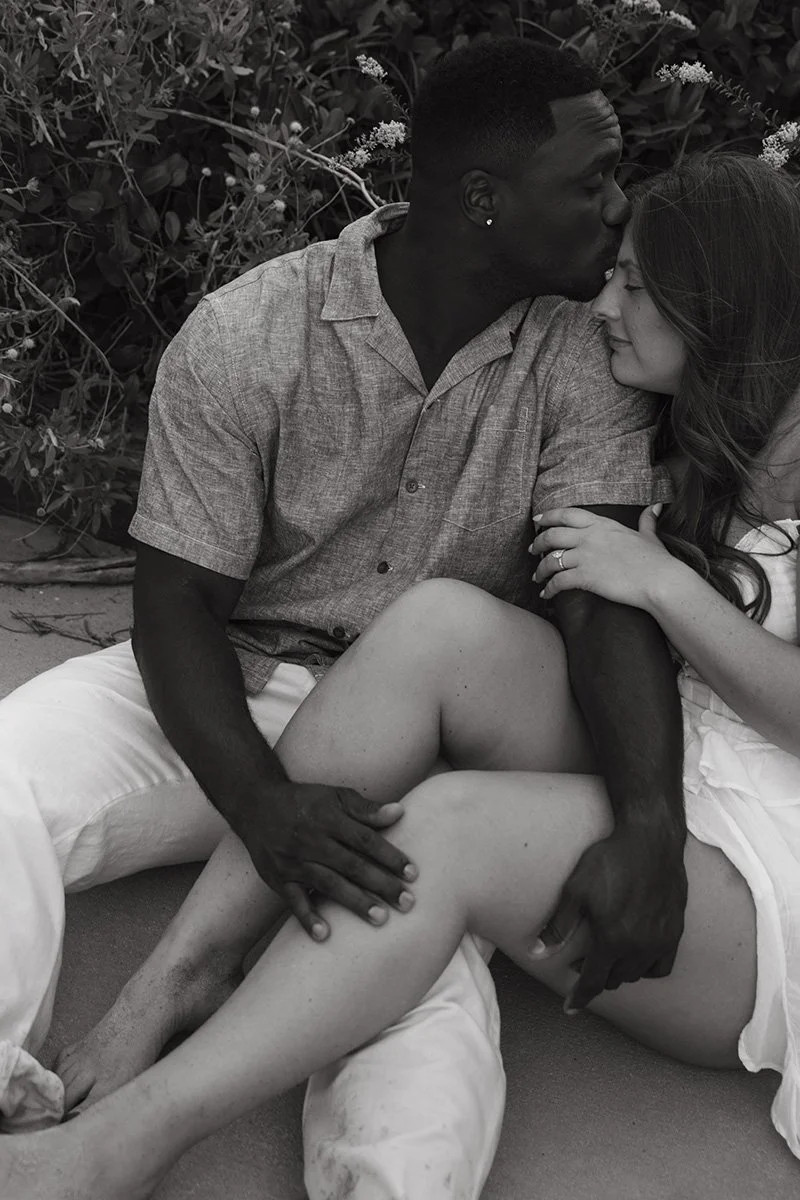 A black man and a white woman sitting closely together outdoors, with the man giving her a gentle kiss on the forehead. The woman is smiling with closed eyes, and the man's hand is resting on her leg. They are surrounded by foliage and flowers.