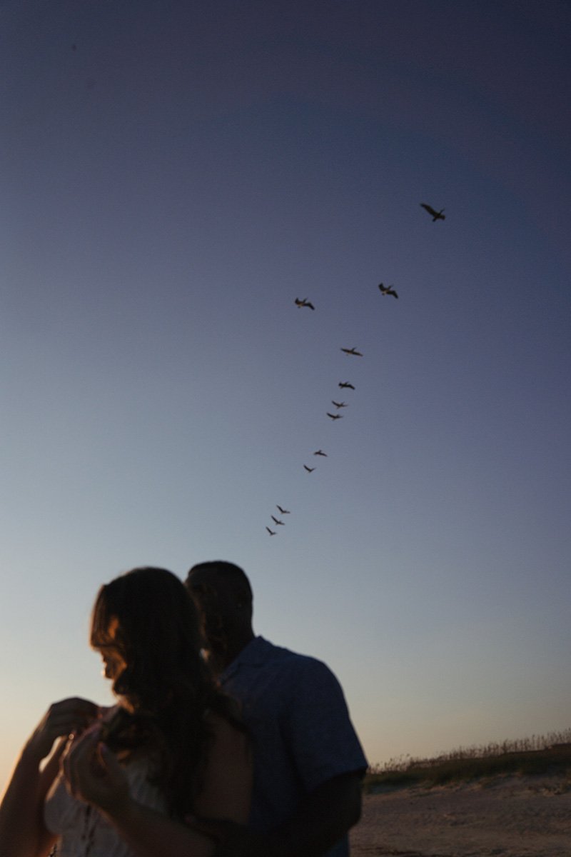 Silhouettes of a couple embracing on a beach at sunset with airplanes flying in a V formation in the sky.