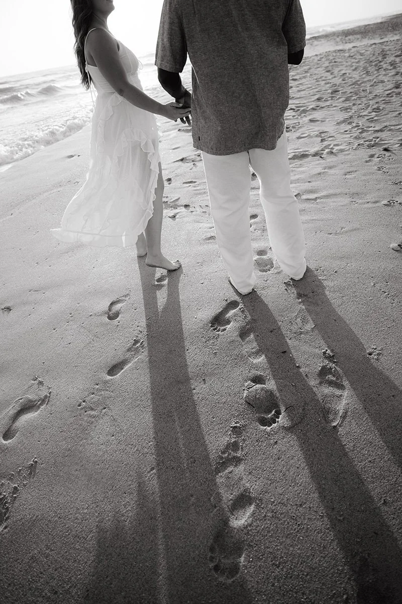 A couple holding hands walking on the beach during sunset, with long shadows cast on the sand.