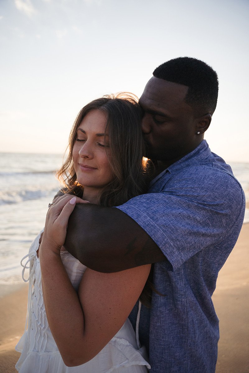 A couple hugging on the beach at sunset, with the ocean in the background.