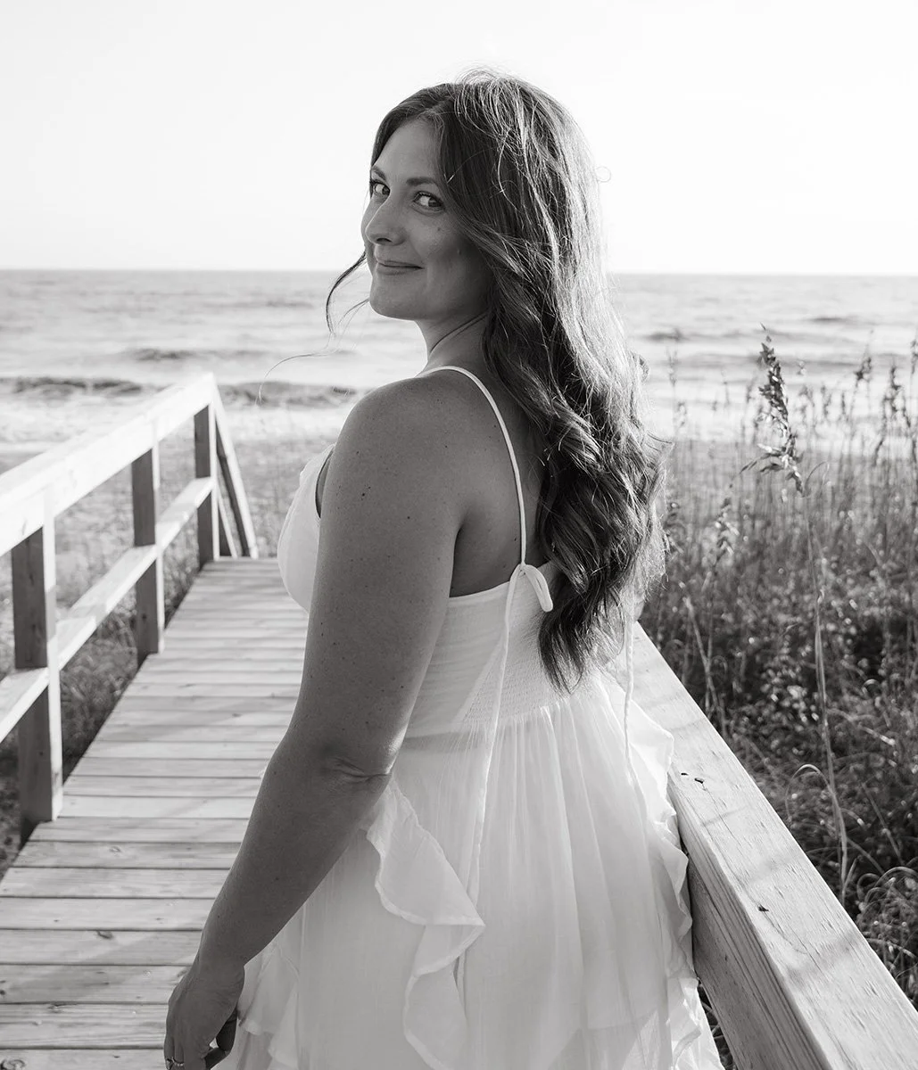 Woman in white dress standing on a wooden pier near the ocean, looking back and smiling.