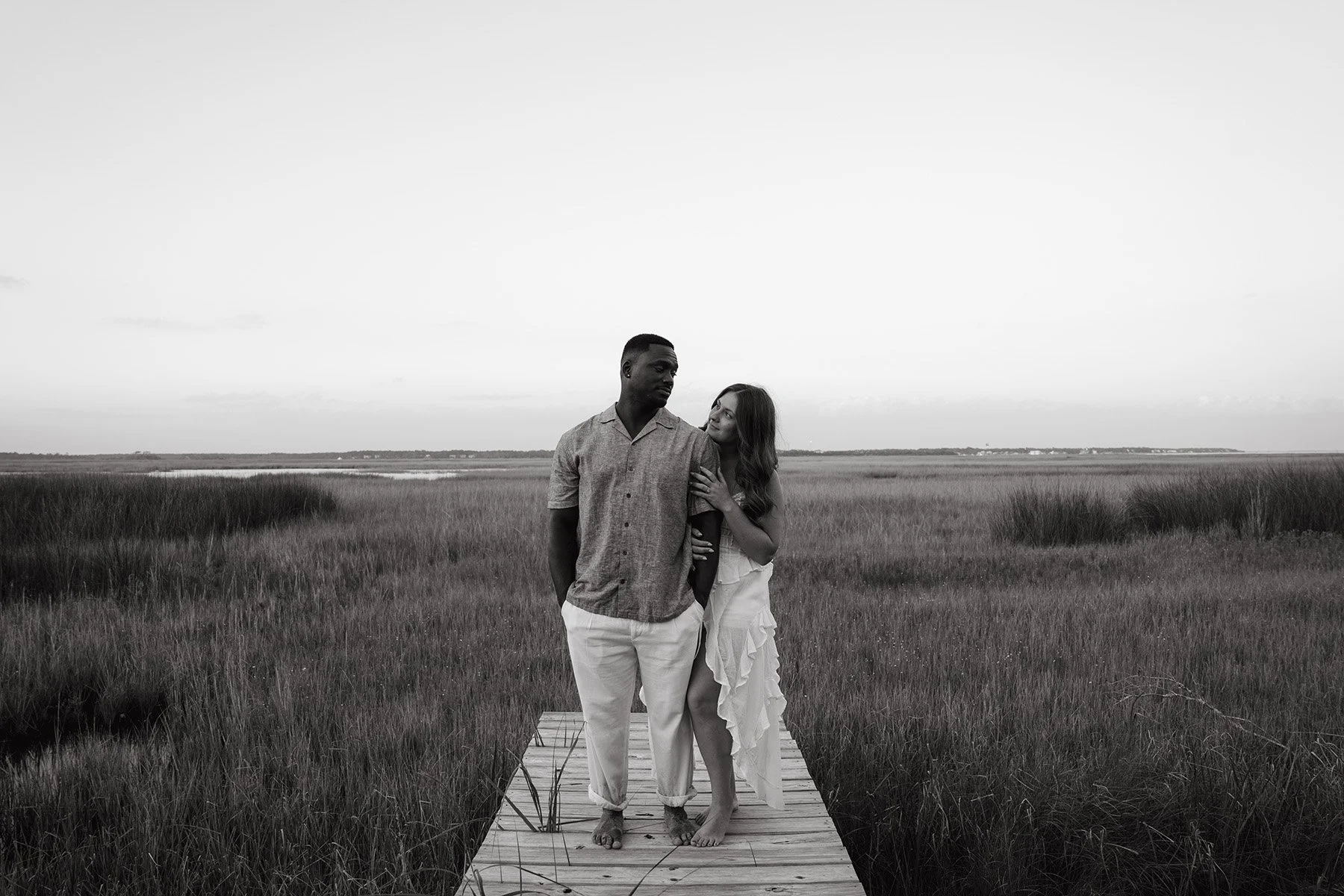 A black and white photo of a couple standing on a wooden dock in a marshland, with tall grass and water in the background, embracing each other lovingly.