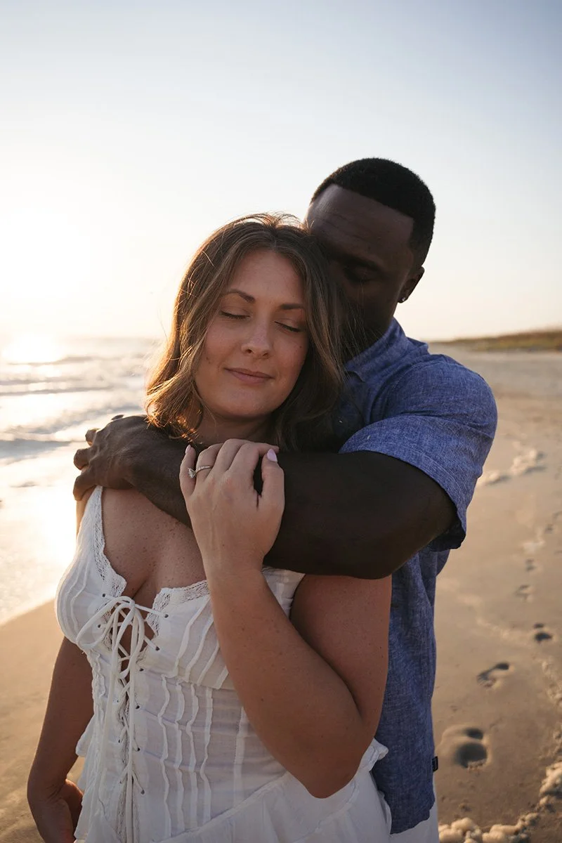 A woman and a man hugging on a beach at sunset, with the ocean and sandy shoreline in the background.