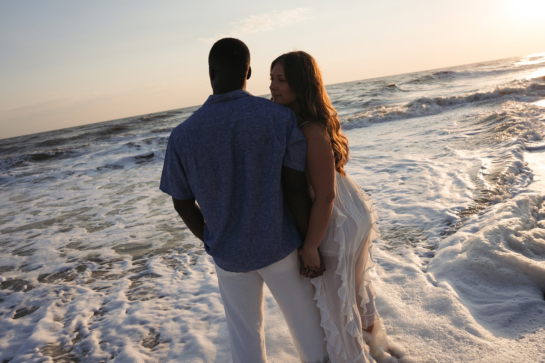 A couple is standing hand in hand on the beach at sunset, with gentle waves and the ocean in the background.