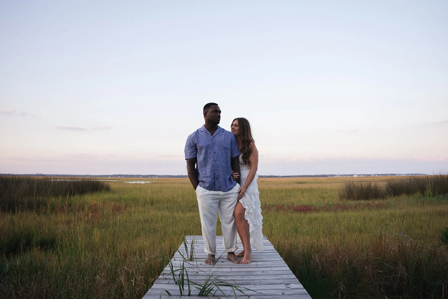A man and woman standing close on a wooden dock in a marsh at sunset, with the woman leaning into the man, both barefoot.