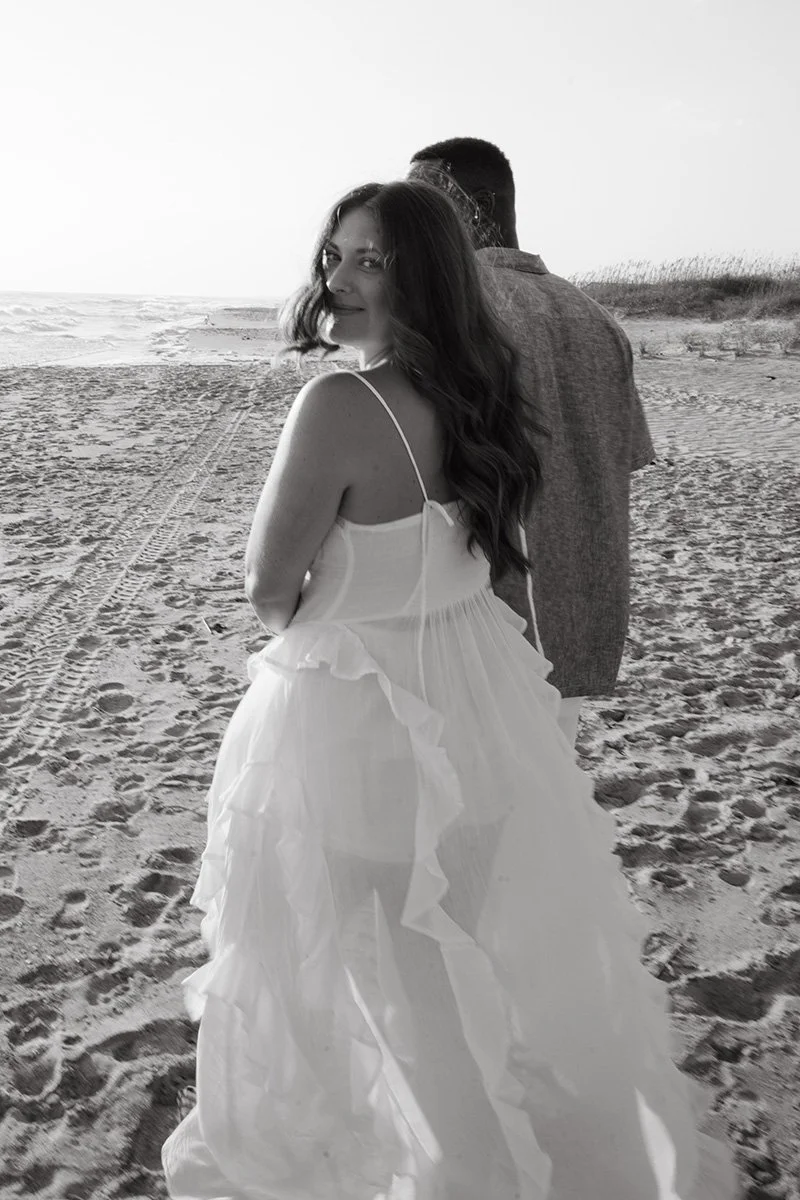 A woman in a white dress smiling at the camera on a sandy beach with a man standing behind her. The photo is in black and white.