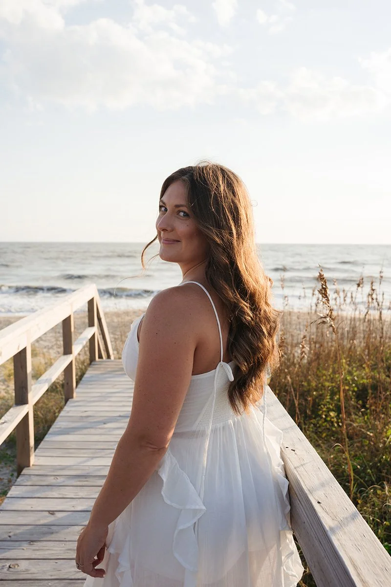 A woman in a white dress standing on a wooden pier by the beach, with ocean waves in the background and a partly cloudy sky.