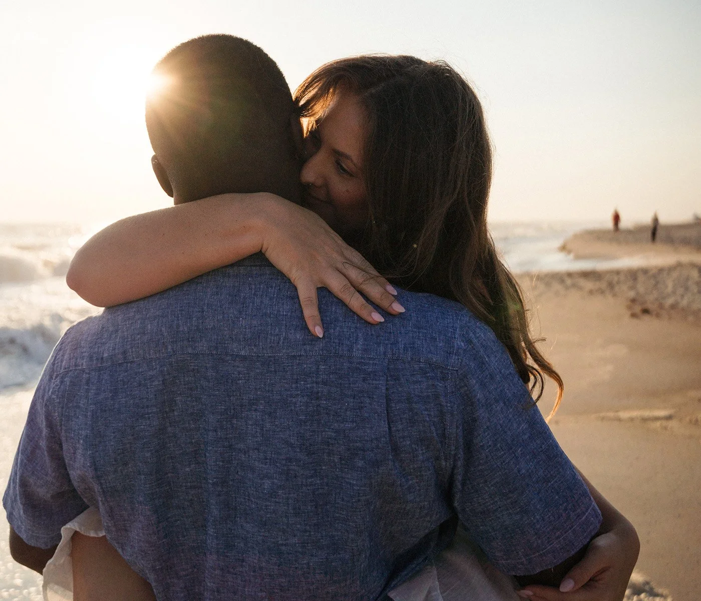 A couple hugging on the beach during sunset, close-up of the woman with long hair smiling and embracing the man, ocean waves in the background.