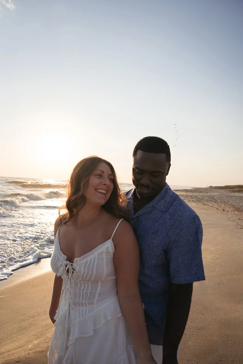 A smiling woman in a white dress standing on a beach at sunset with a man in a blue shirt.