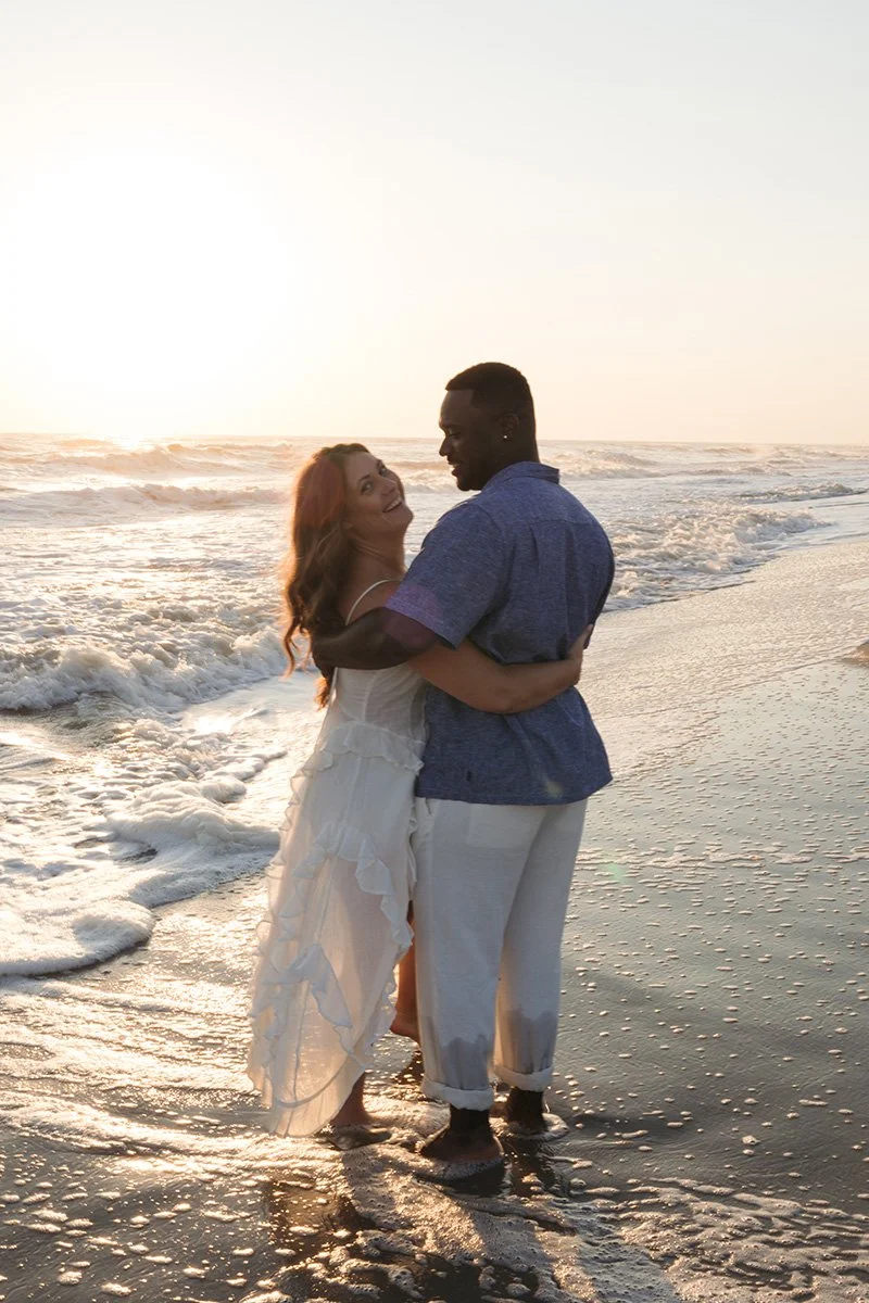A couple hugging on the beach at sunset, with waves in the background.