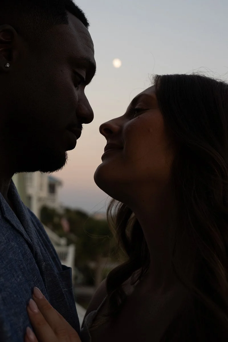 A close-up silhouette of a man and woman facing each other with their foreheads almost touching, outdoors during sunset with the moon visible in the background.