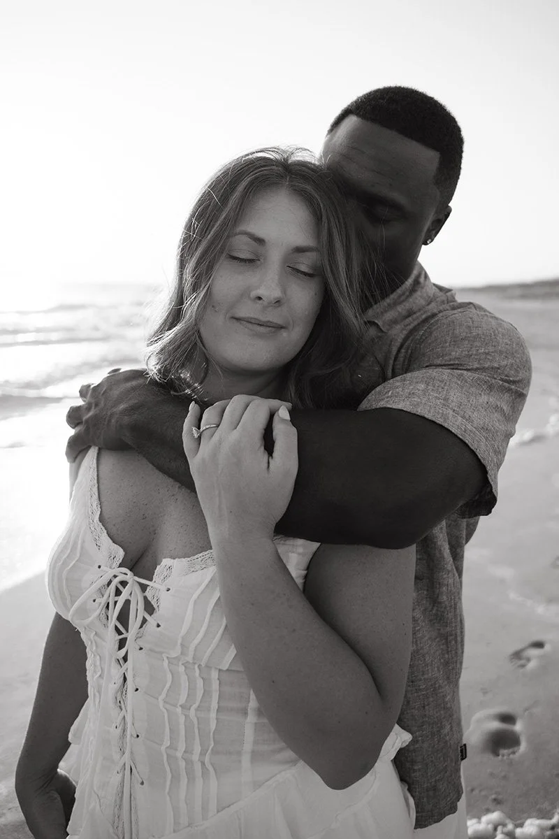 A black-and-white photo of a couple embracing on the beach, with the woman smiling with eyes closed and the man hugging her from behind.