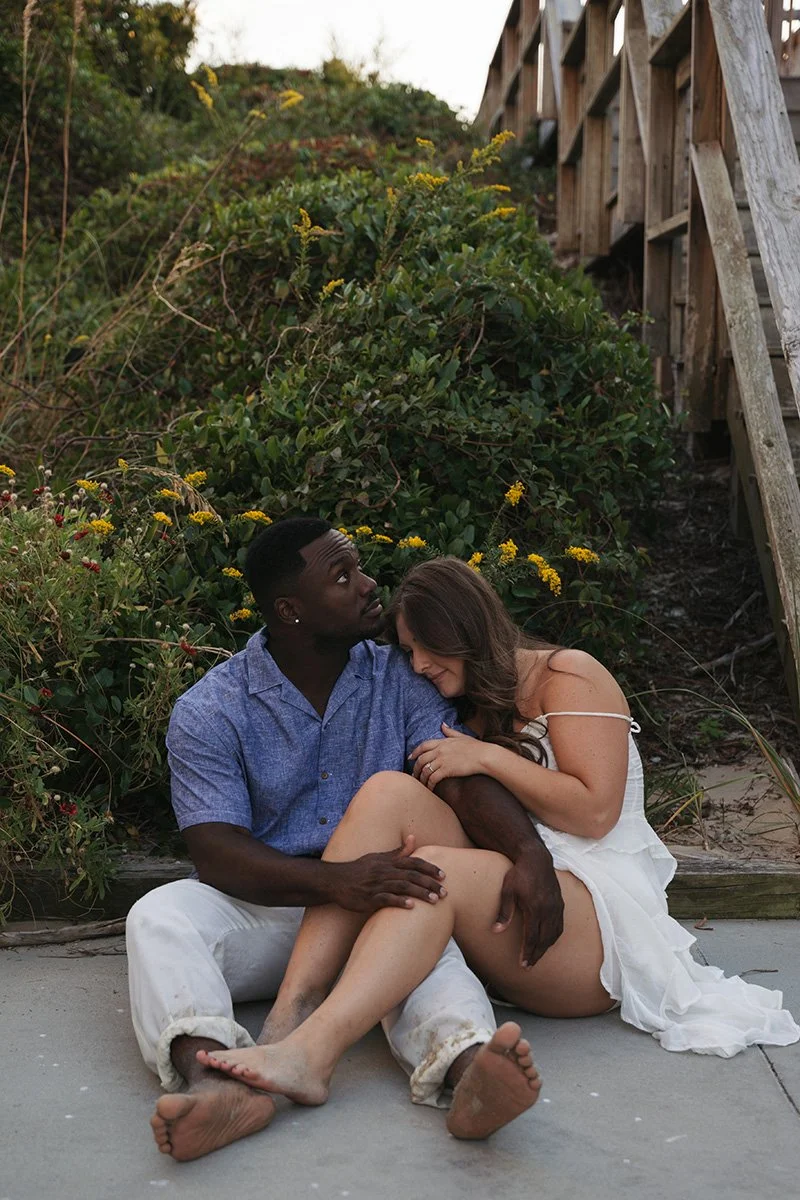 A couple sitting on the sand by flowers and greenery, embracing each other affectionately under a wooden staircase.