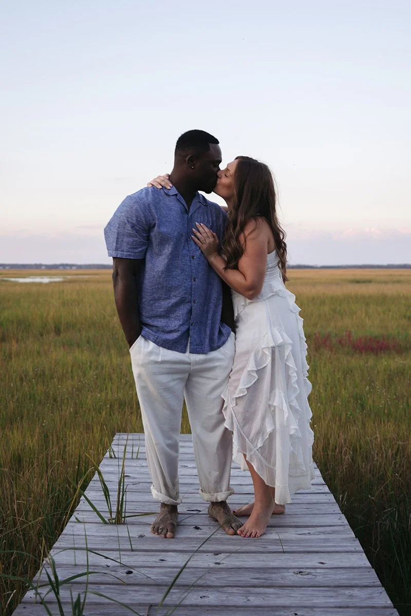 A couple kissing, standing on a wooden dock in a grassy field with a sunset sky in the background.