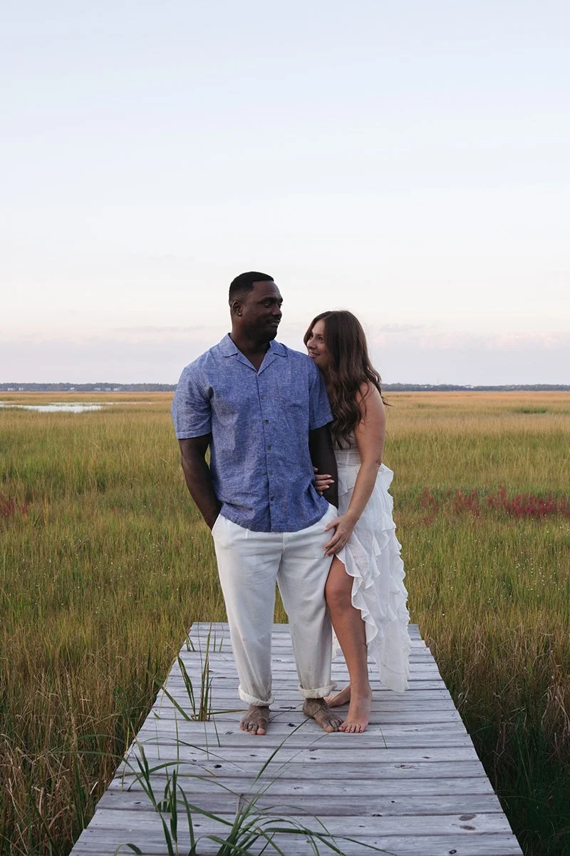 A couple standing on a wooden dock in a marsh at sunset, looking at each other, with a vast sky and marshland in the background.