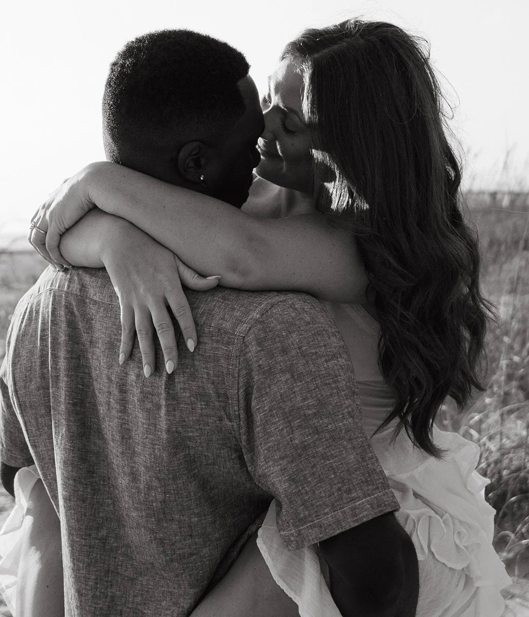 A black-and-white photo of a couple embracing, with the woman hugging the man from the front and their faces close together, outdoors with a blurred background.