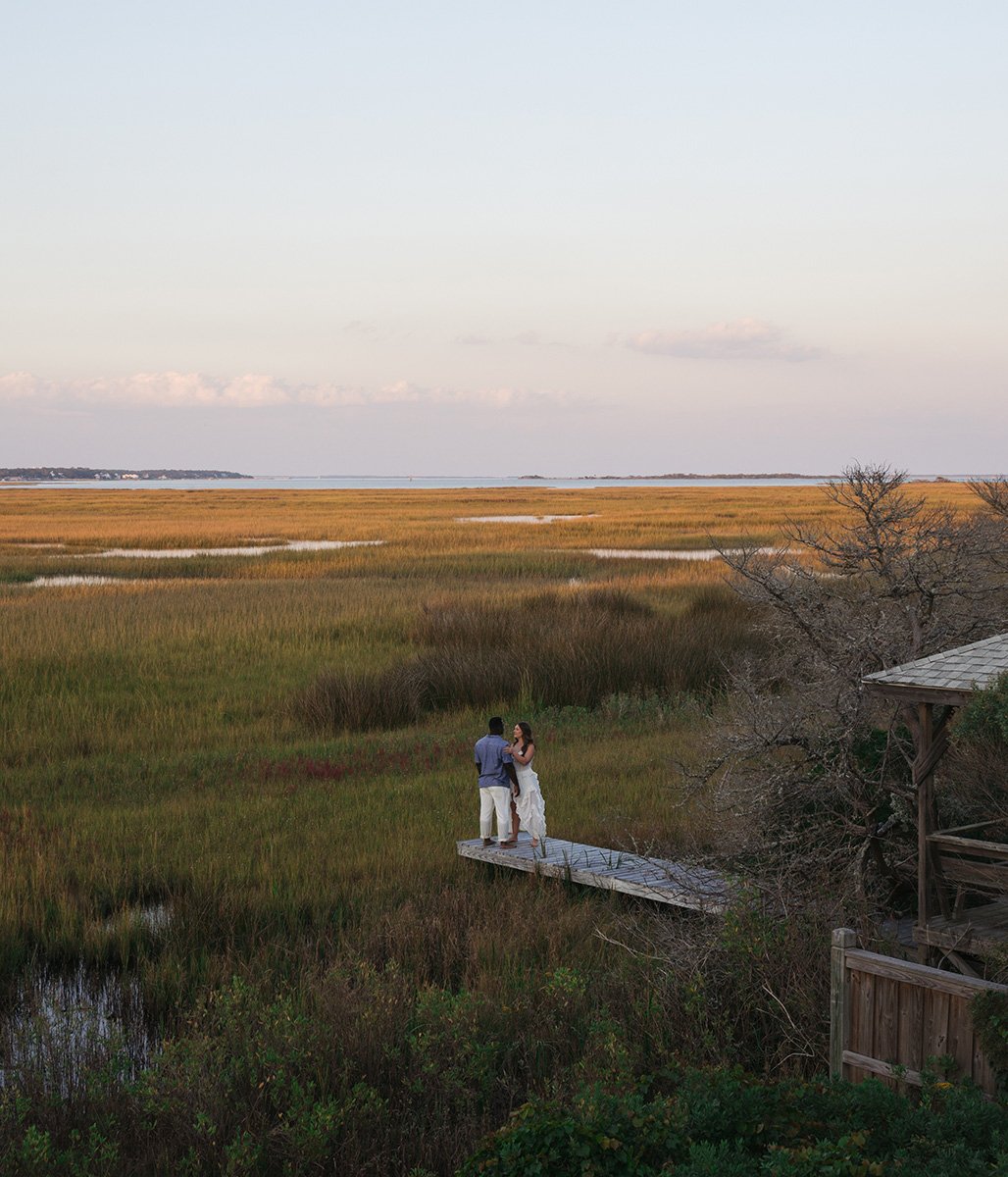 A couple stands on a wooden dock by marshland with tall grasses, near a body of water, under a mostly clear sky during sunset or sunrise.