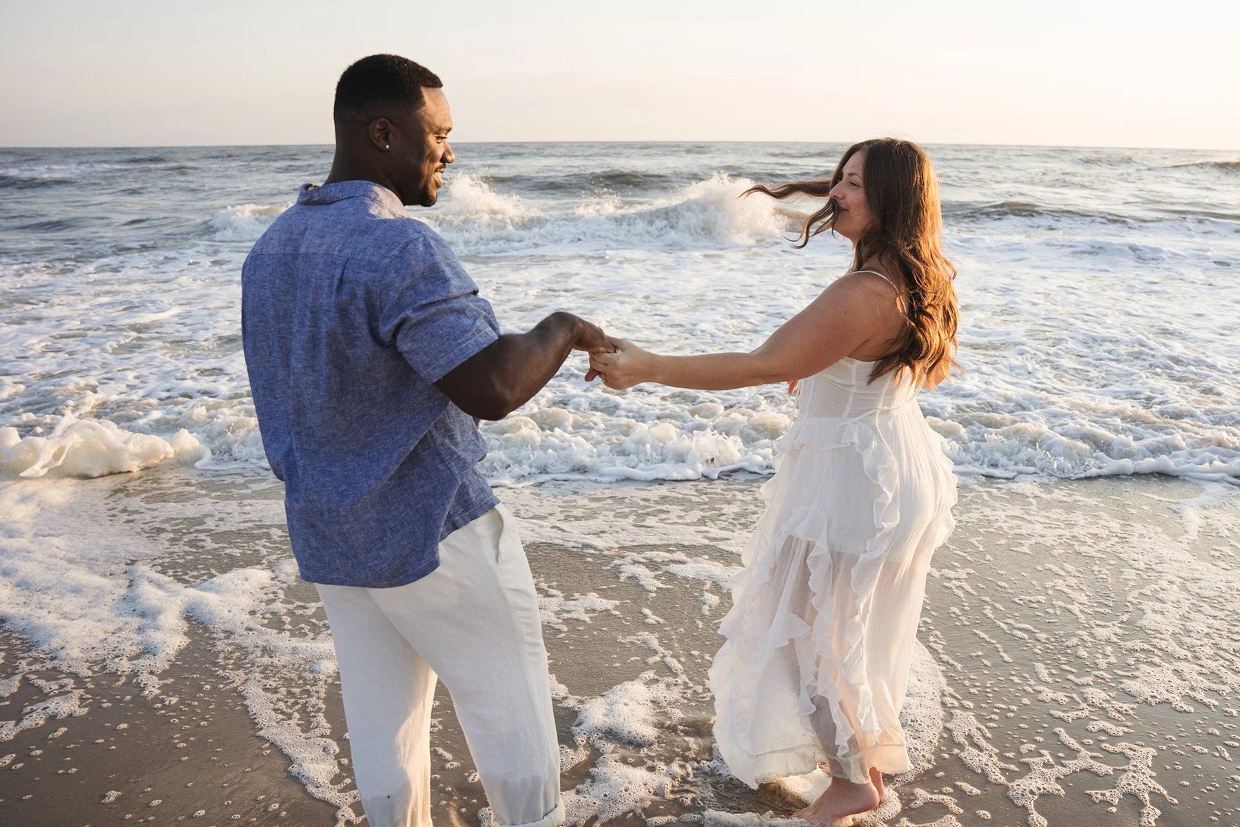 A couple is holding hands and dancing on the shoreline of the ocean during sunset. The woman is wearing a white dress, and the man is wearing a blue shirt and white pants.
