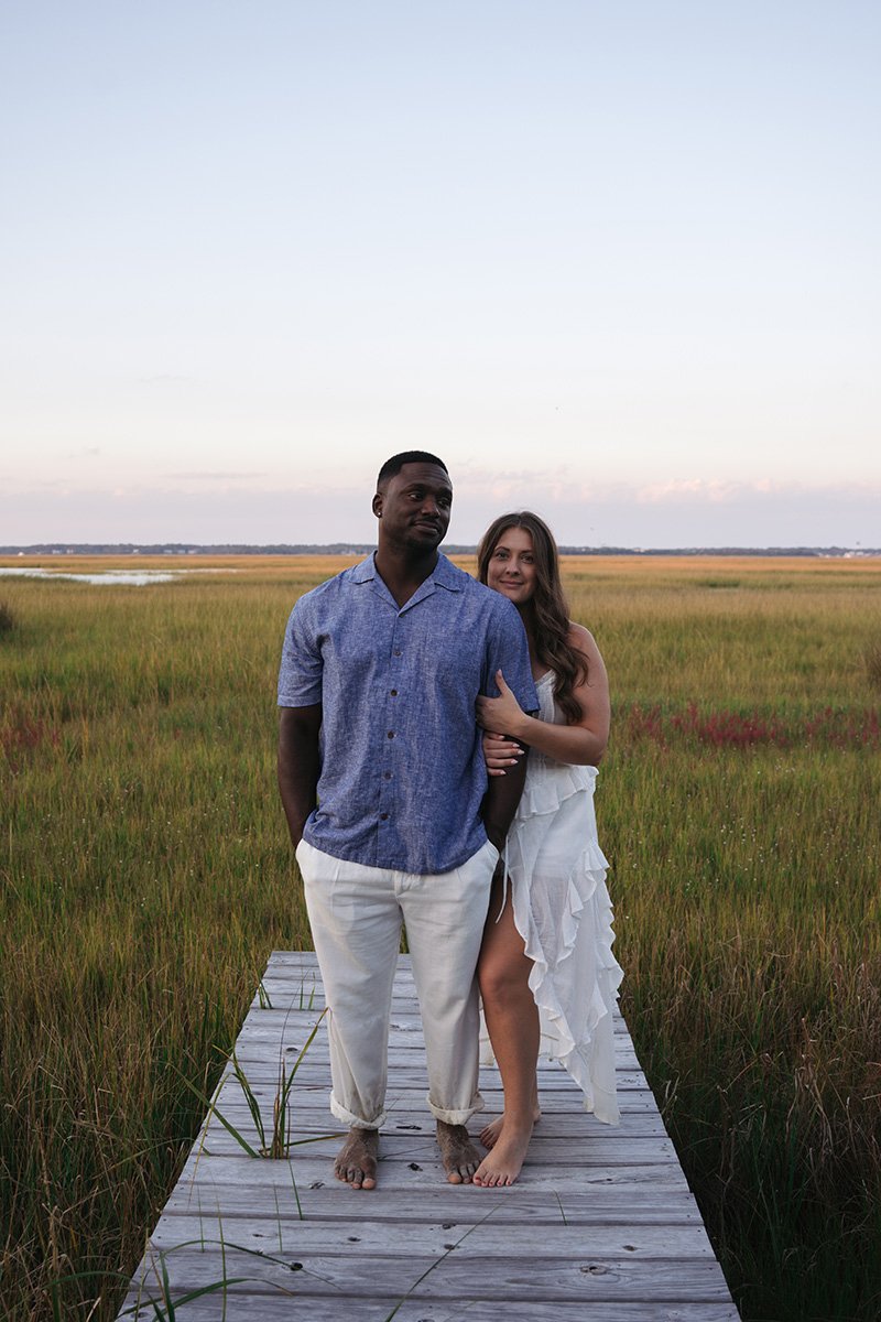 A couple stands on a wooden dock in a marshland at sunset, with green grass and a body of water in the background.