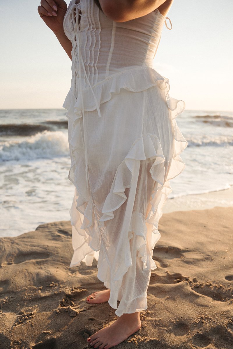 A person in a flowing, ruffled white dress standing barefoot on the sandy beach near the ocean during sunset.