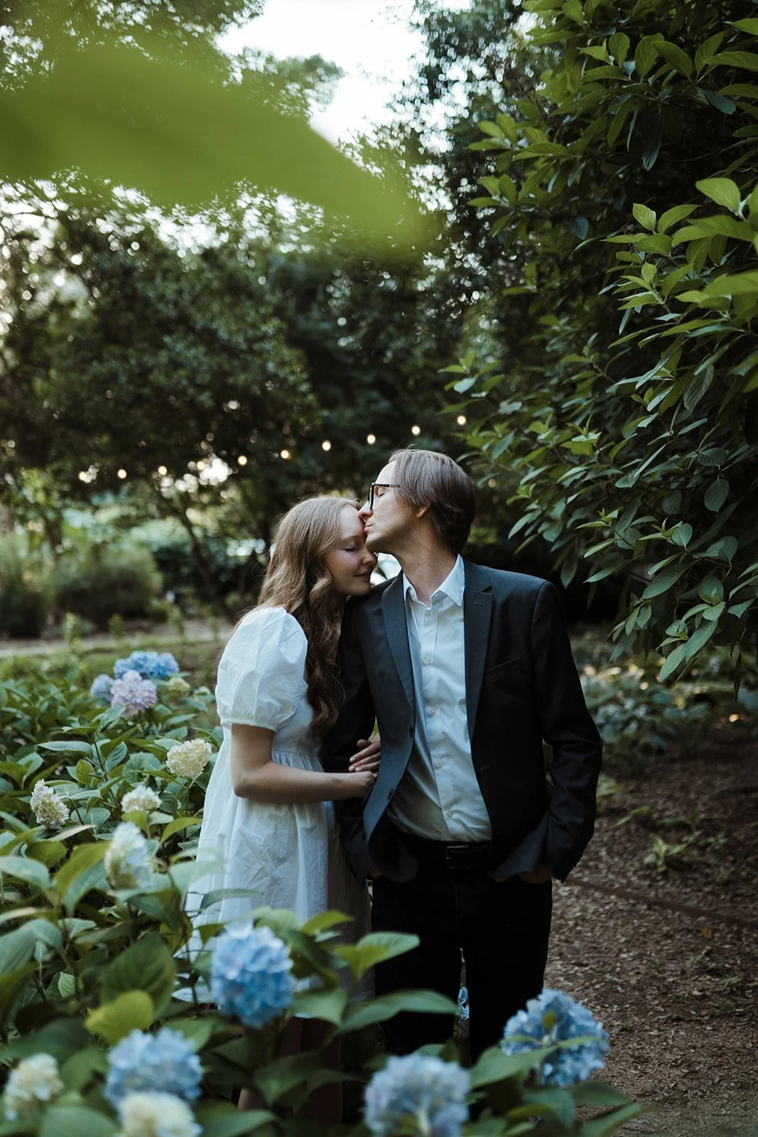 This is an image of a couple having their photo taken at the McGill Rose Garden in Charlotte, North Carolina. They are standing, cuddling, with their eyes closed and touching cheeks. There are large blue flowers and green bushes surrounding them.