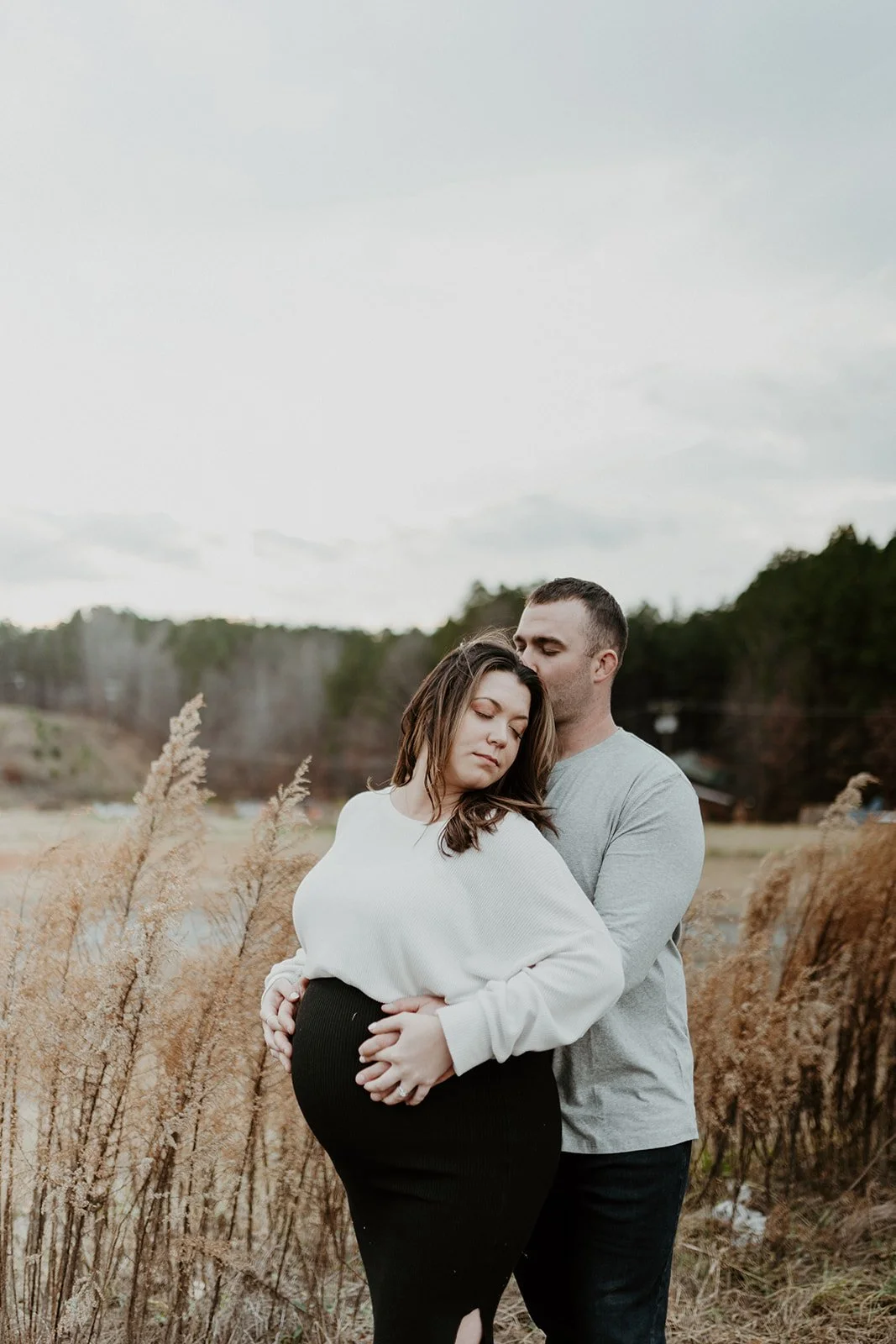 This is an image of a couple embracing during their maternity session at the White Water Center in Charlotte, North Carolina. The woman is visibly pregnant, holding her belly with her eyes closed. The father stands behind her holding her, kissing her