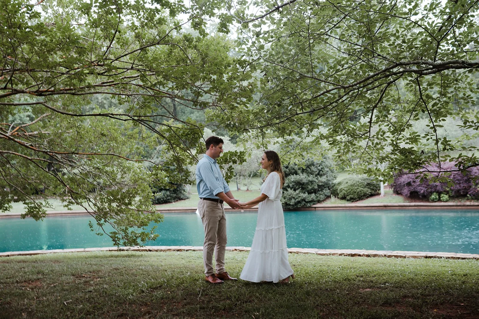 This is an image of a couple having their photo taken at Freedom Park in Charlotte, North Carolina. they are standing under large trees, holding hands in front of a pond.