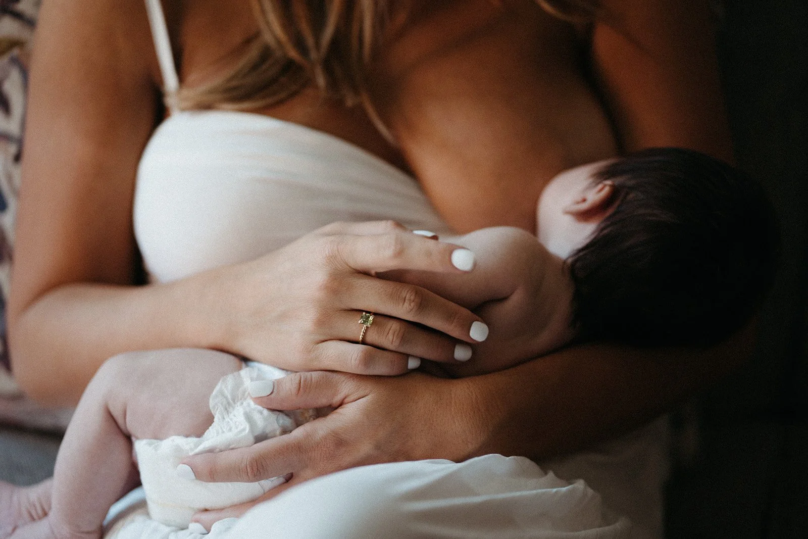 A mother sits in a white dress breastfeeding her infant. She is gently holding her baby with her wedding ring visible.