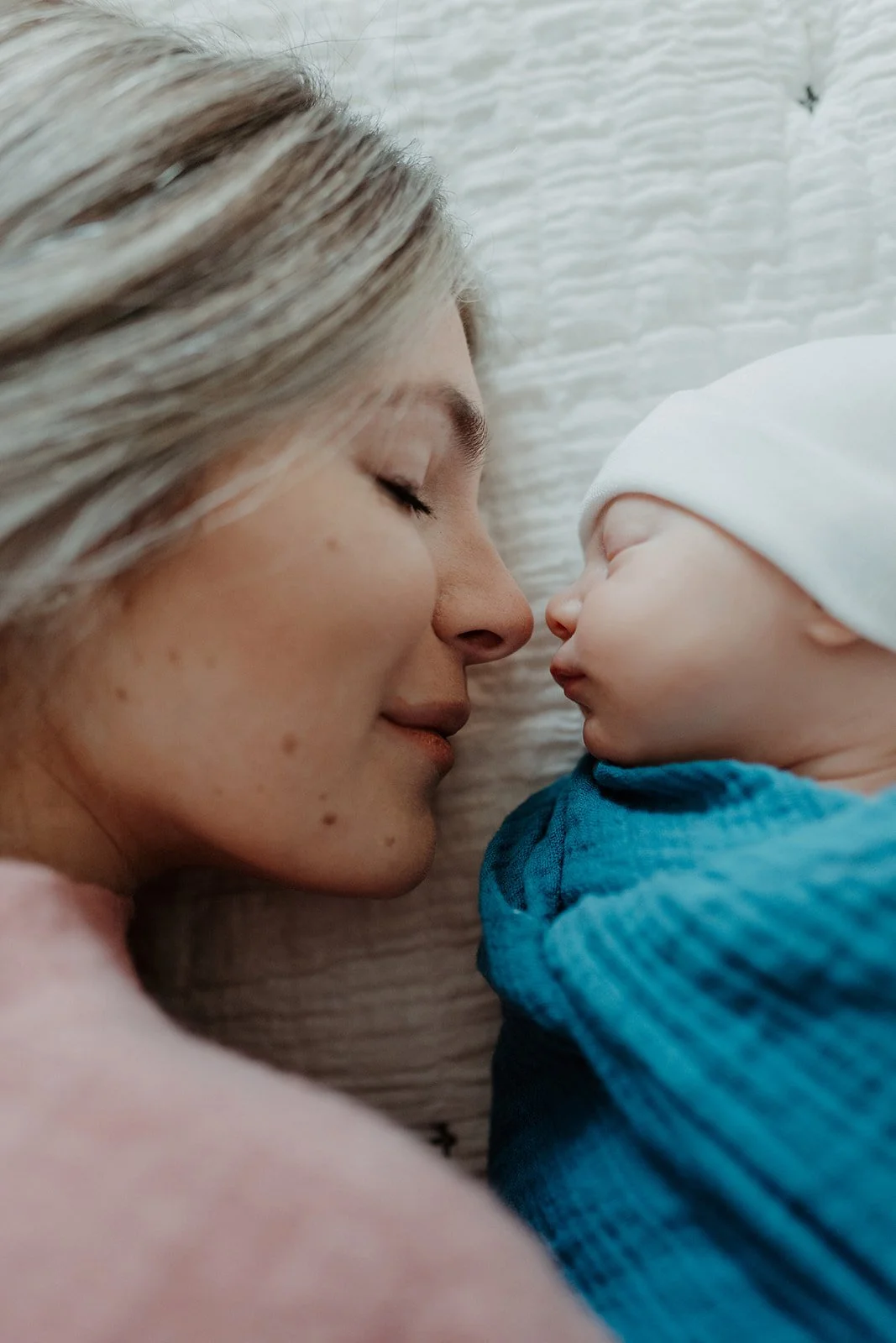 A woman lays on a linen blanket, nose to nose with her infant child during a newborn session. Both their eyes are closed.