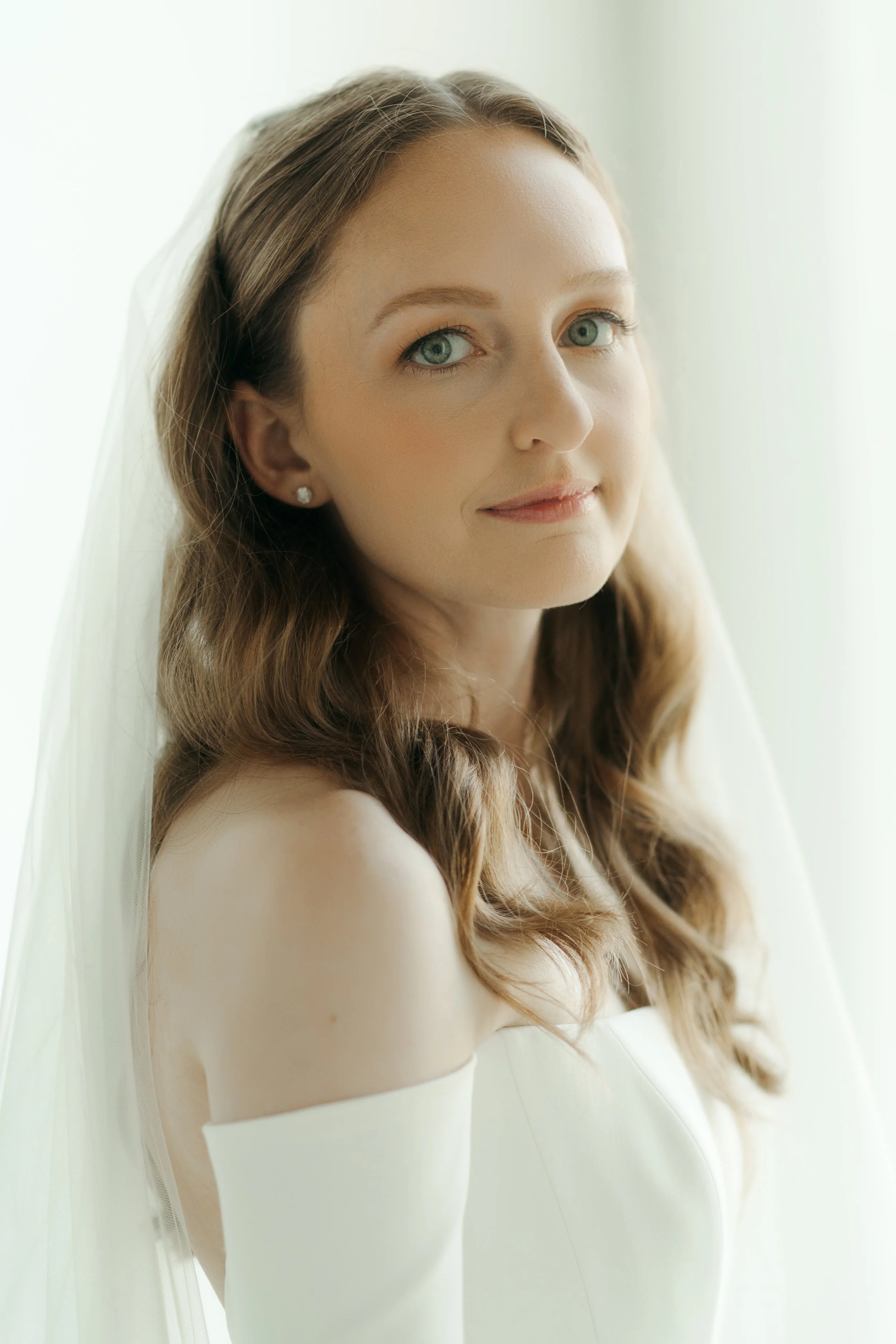 A woman stares at the camera in an all white room wearing her wedding gown and veil for bridal portraits