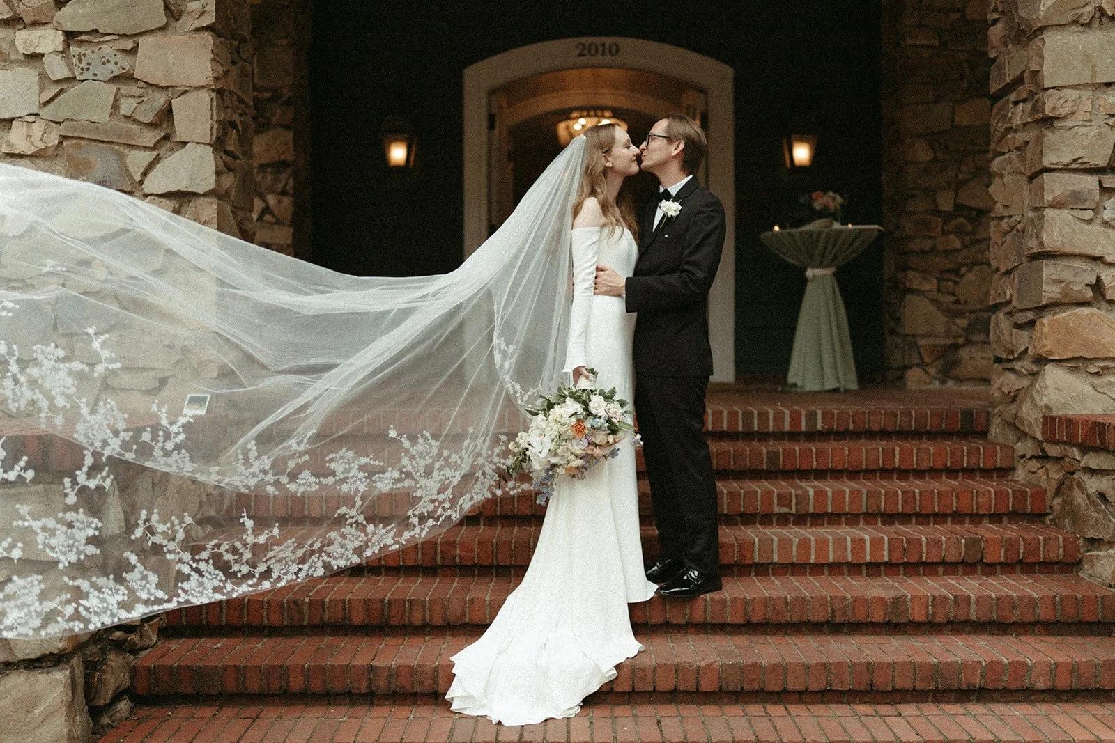 A couple in a white dress and black tux stands kissing on the steps of the VanLandingham Estate during their wedding day.