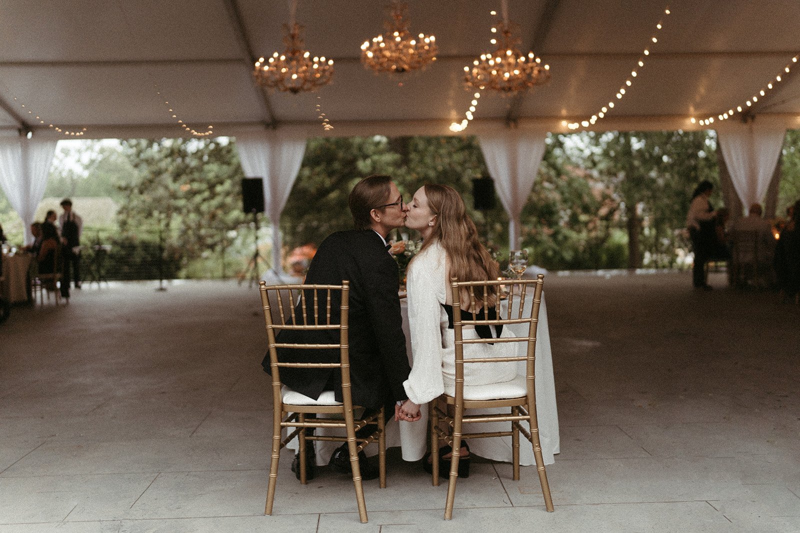  A couple kisses in wooden chairs under glowing chandeliers while seated at their wedding at the VanLandingham estate. 