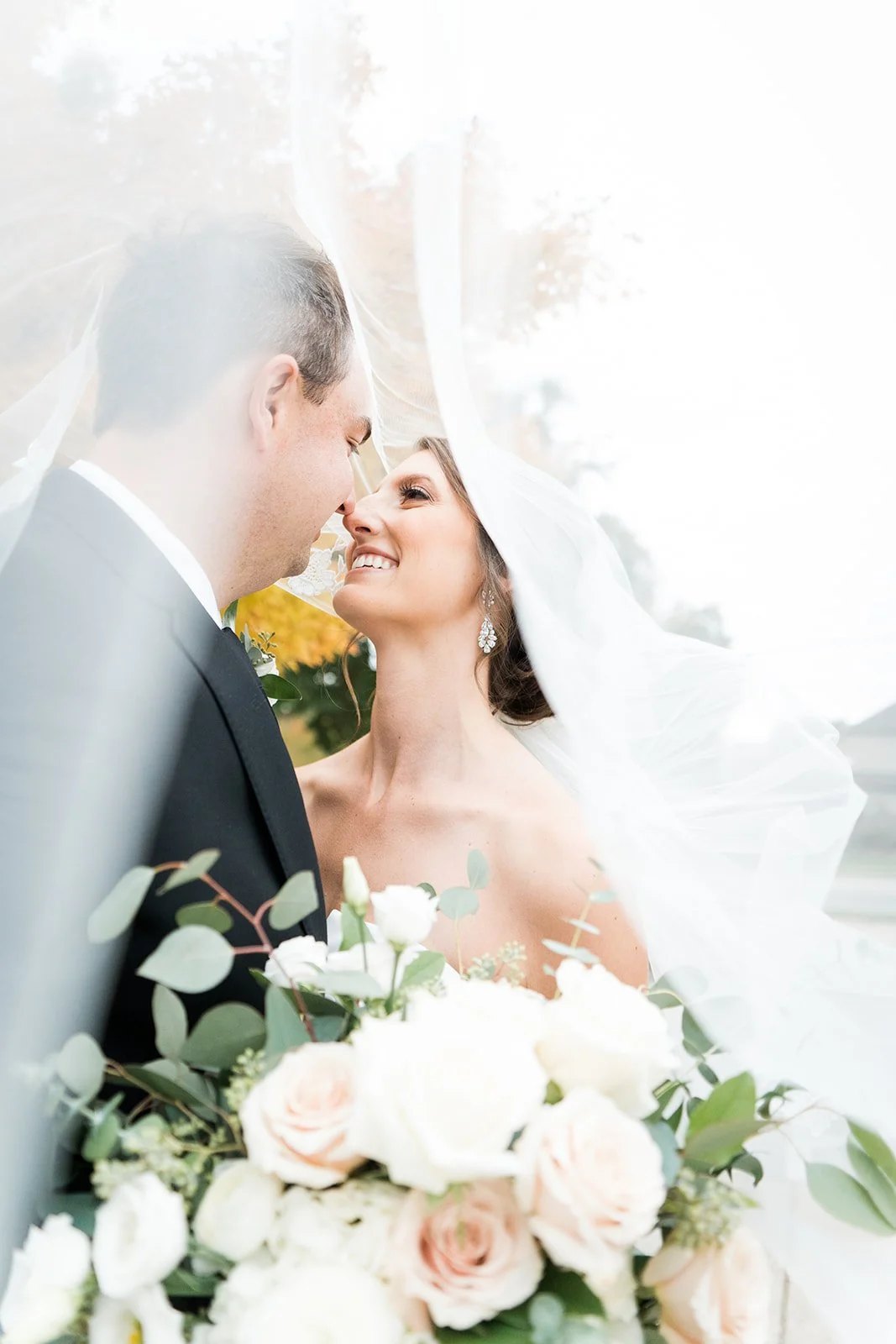 A bride and groom stand nose to nose smiling under a veil on their wedding day.
