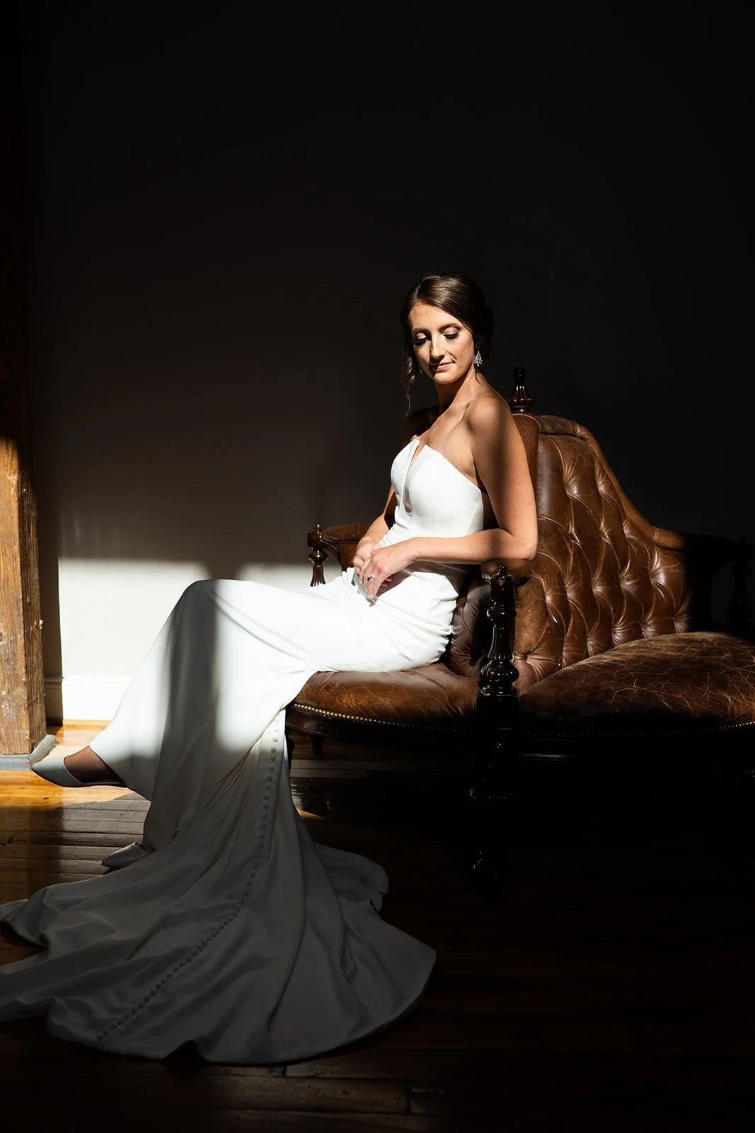 A woman sits in her wedding dress on a leather chair in dramatic, high contrast light.
