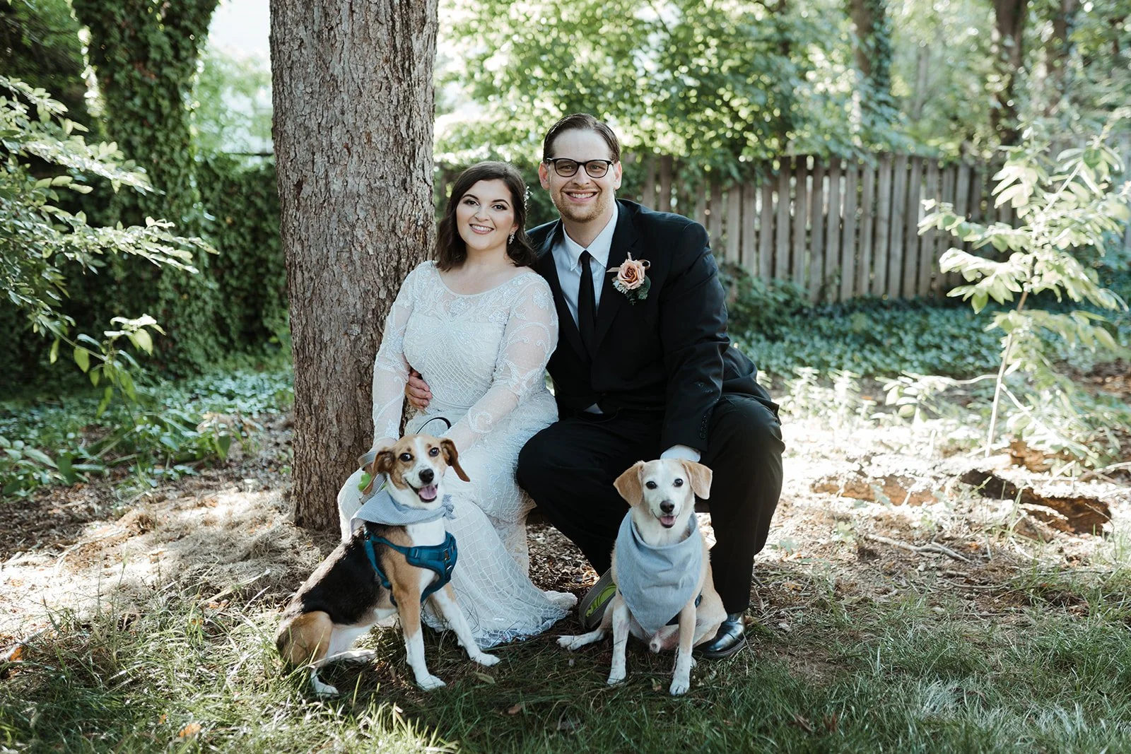 A couple squats down on their wedding day to pose with their two small dogs. They are all facing the camera and smiling.