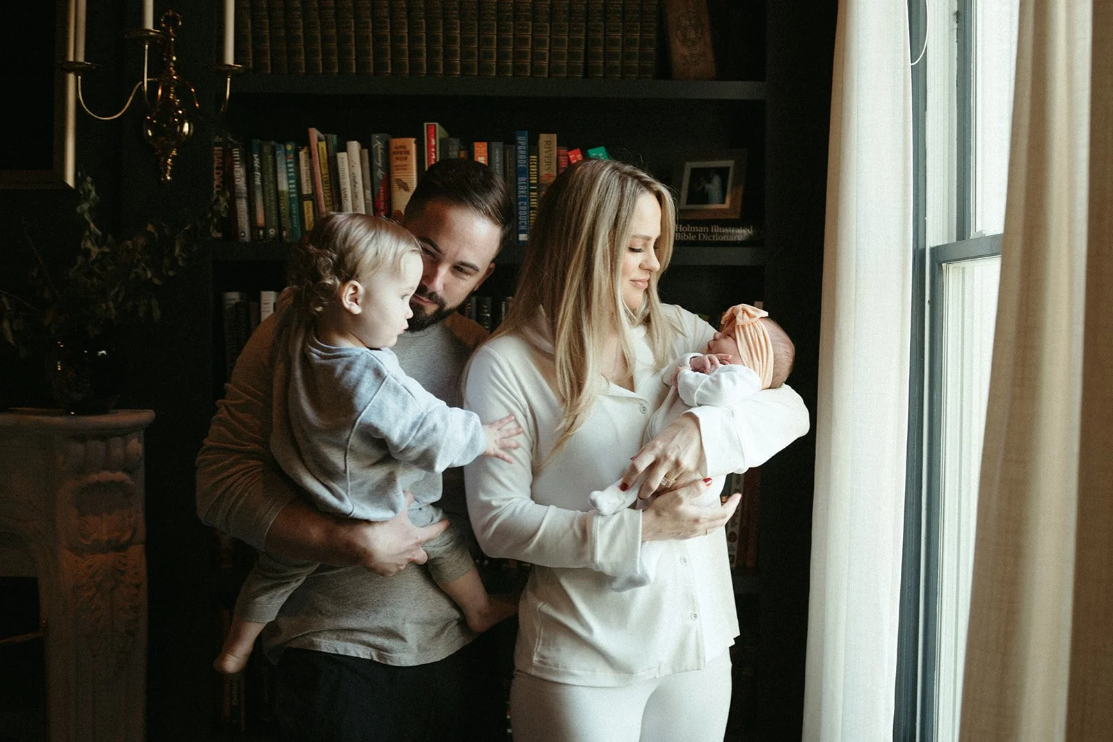 A young couple stands by a window in their home holding their newborn and toddler for an at home newborn photo session.