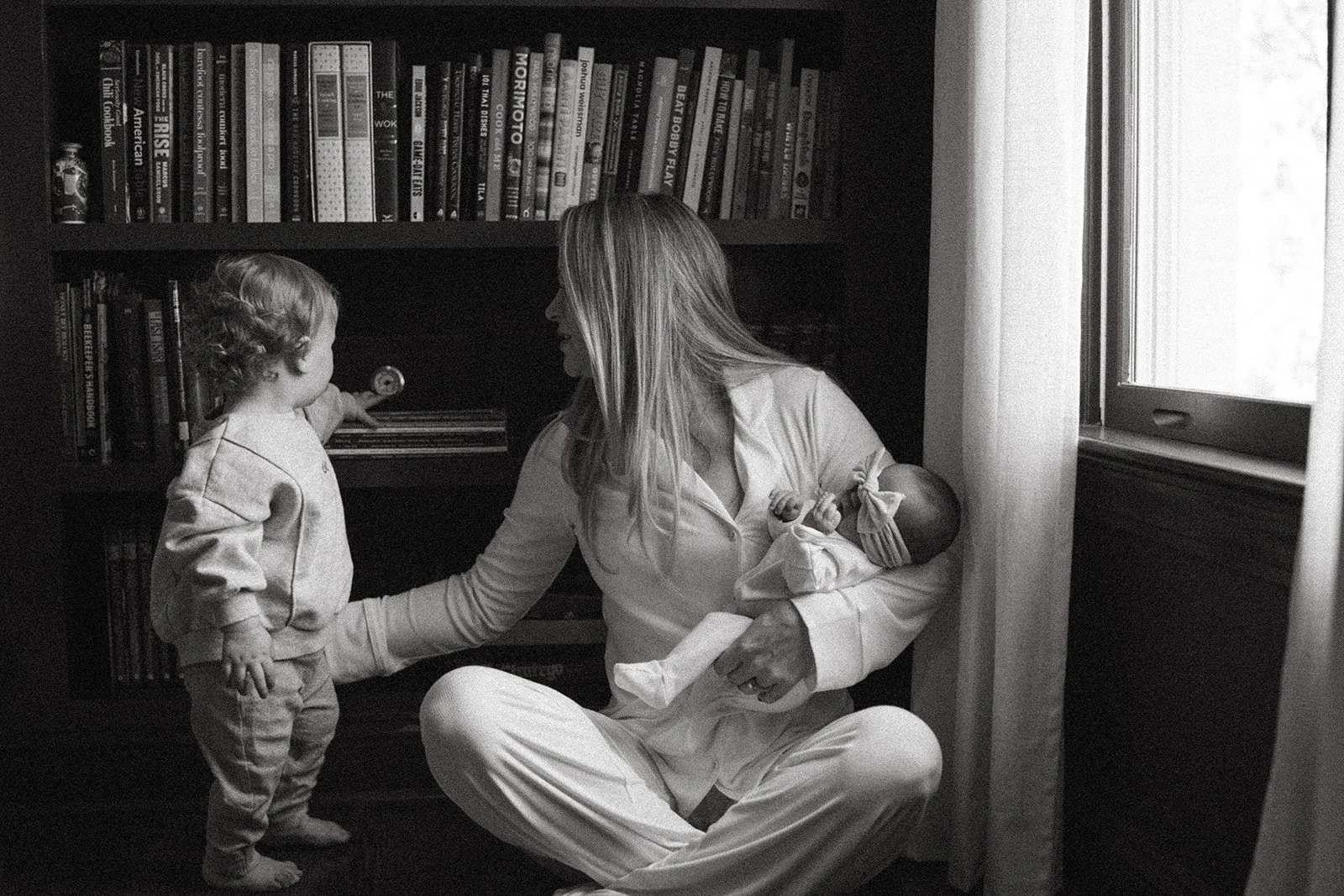 A mother sits cross legged on the floor holding her newborn in one arm and holding up her toddler with the other in front of a bookshelf