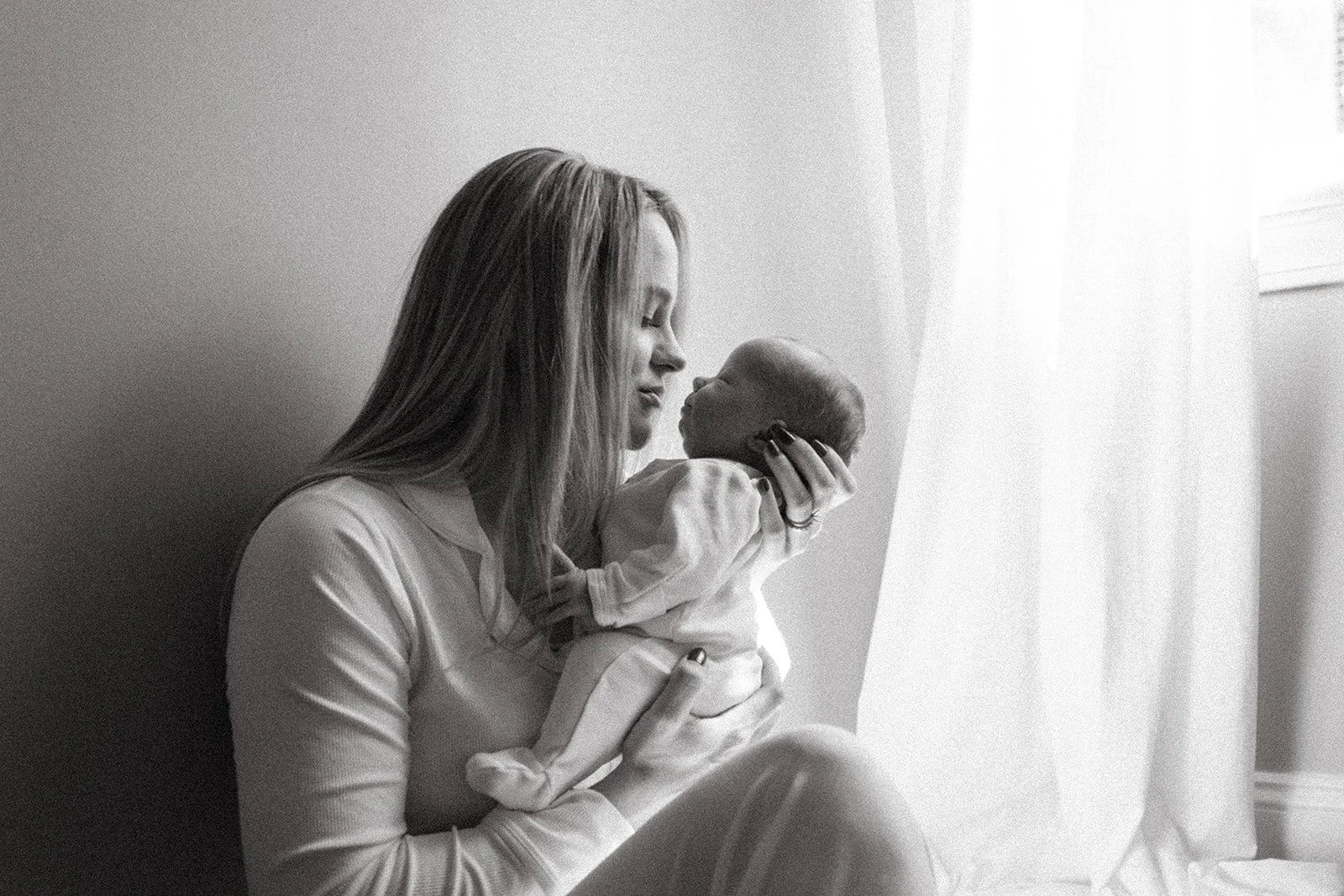 A mother cradles her newborn to her nose near a bright window for an at home newborn photoshoot