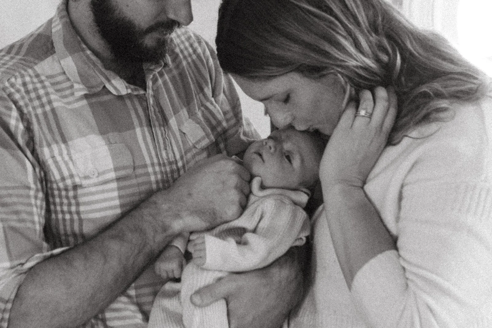 New parents hold their infant during their newborn photo session. The father is cradling the baby in his arm and the mother is leaning down to kiss the baby on the forehead. The image is black and white.
