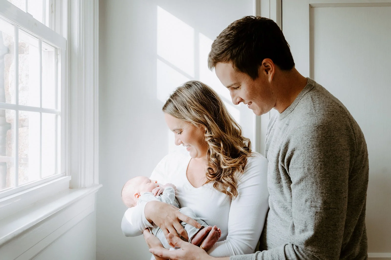 This is a photo of a heterosexual couple holding their infant baby during their newborn photography photo session. They are standing near a bright window holding and looking at their sleeping baby.
