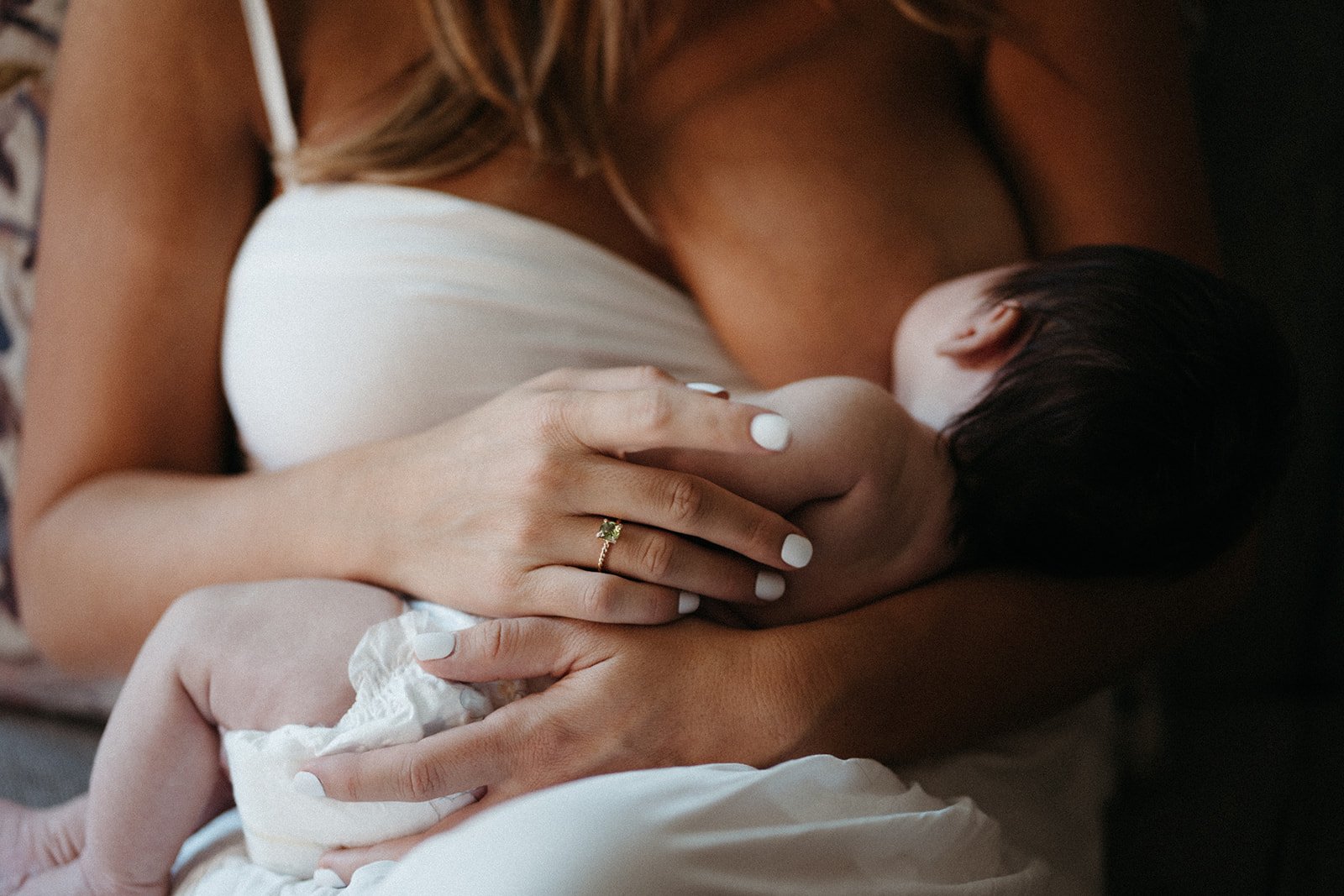  This is an image of a woman breastfeeding her infant during a newborn photo session. She is visible from the neck to the waist, sitting. She is holding her infant in her arms as the baby feeds. 