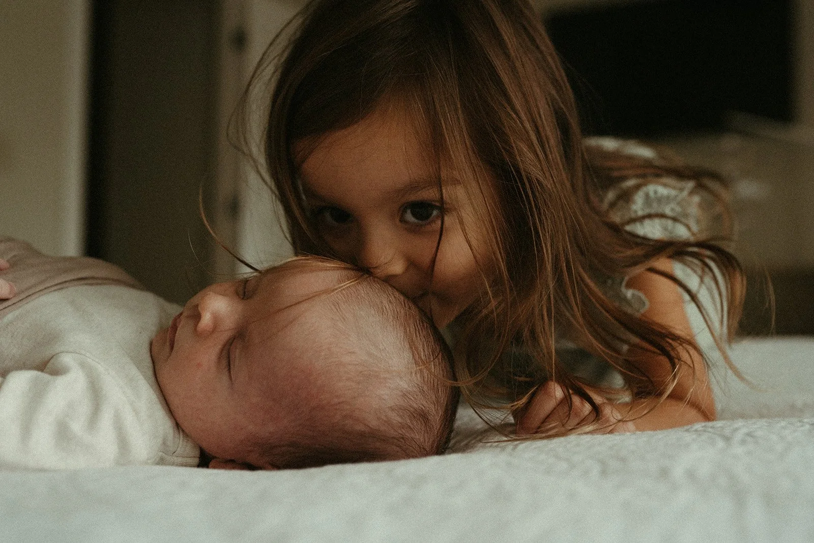  This is an image of an infant lying on a bed during a newborn photo shoot. An older female sibling is leaning over kissing the baby’s forehead. 