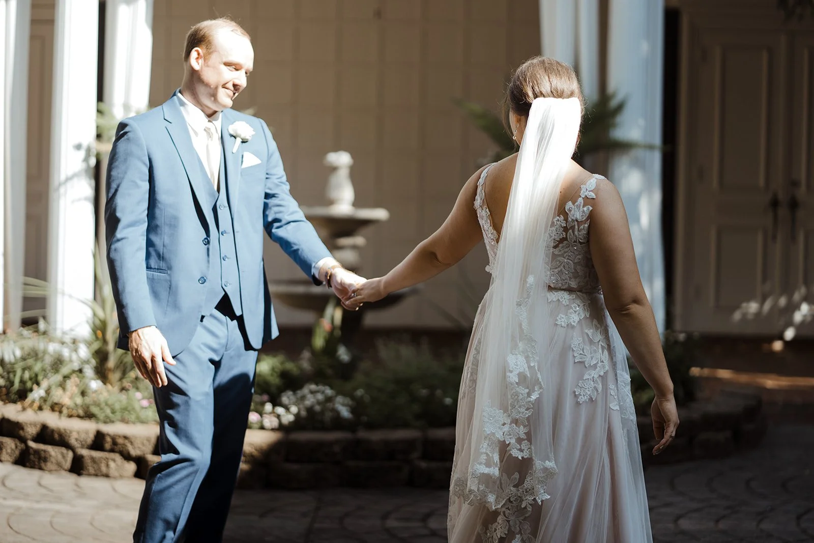 A wedding couple stands outside in front of a fountain holding hands during their wedding day first look.