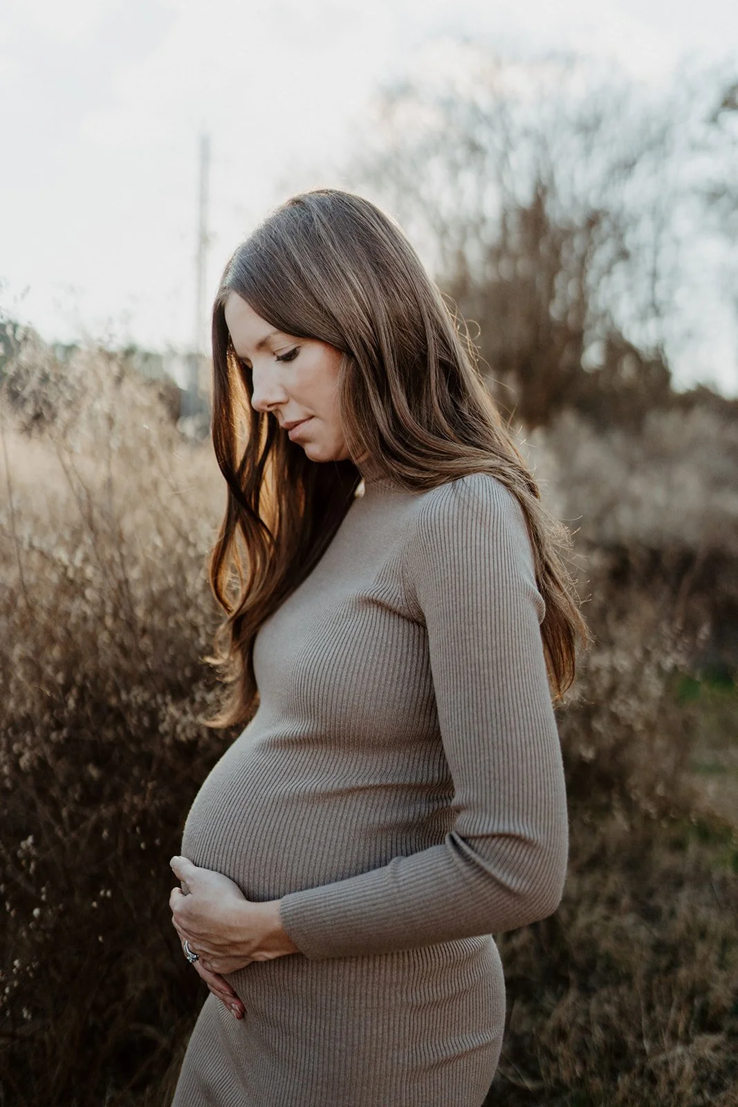 This is a photo of a woman holding her pregnant belly during her maternity shoot. She is wearing a long, knit sweater dress and looking down. She is standing in a field and the sun is shining bright behind her, through her brown hair.