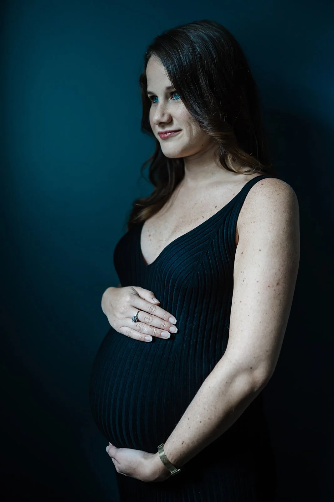 This is an image of a light skinned pregnant woman holding her belly during her maternity photo shoot. She is wearing a sleeveless, knit black dress. She is standing near a dark blue backdrop, and looking to the left of the frame.