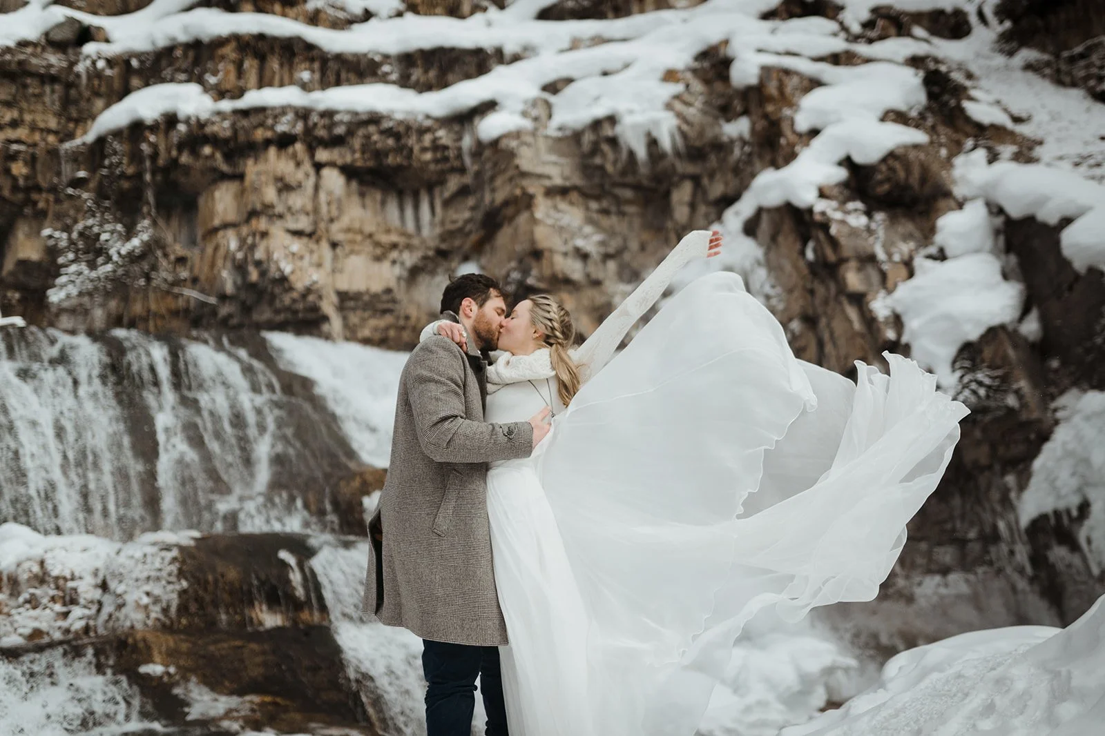A couple stands near a frozen waterfall during their winter elopement. They are kissing and the woman is throwing her long white dress behind her in the wind.