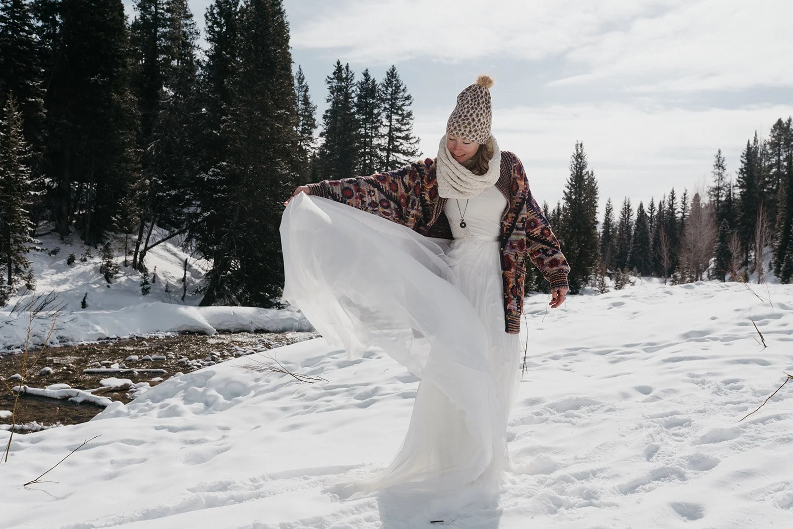 A woman stands in the snow on a bright day wearing a dress and warm winter clothes for her winter elopement. She is wearing a knit hat, scarf and sweater and holding the skirt of her dress.