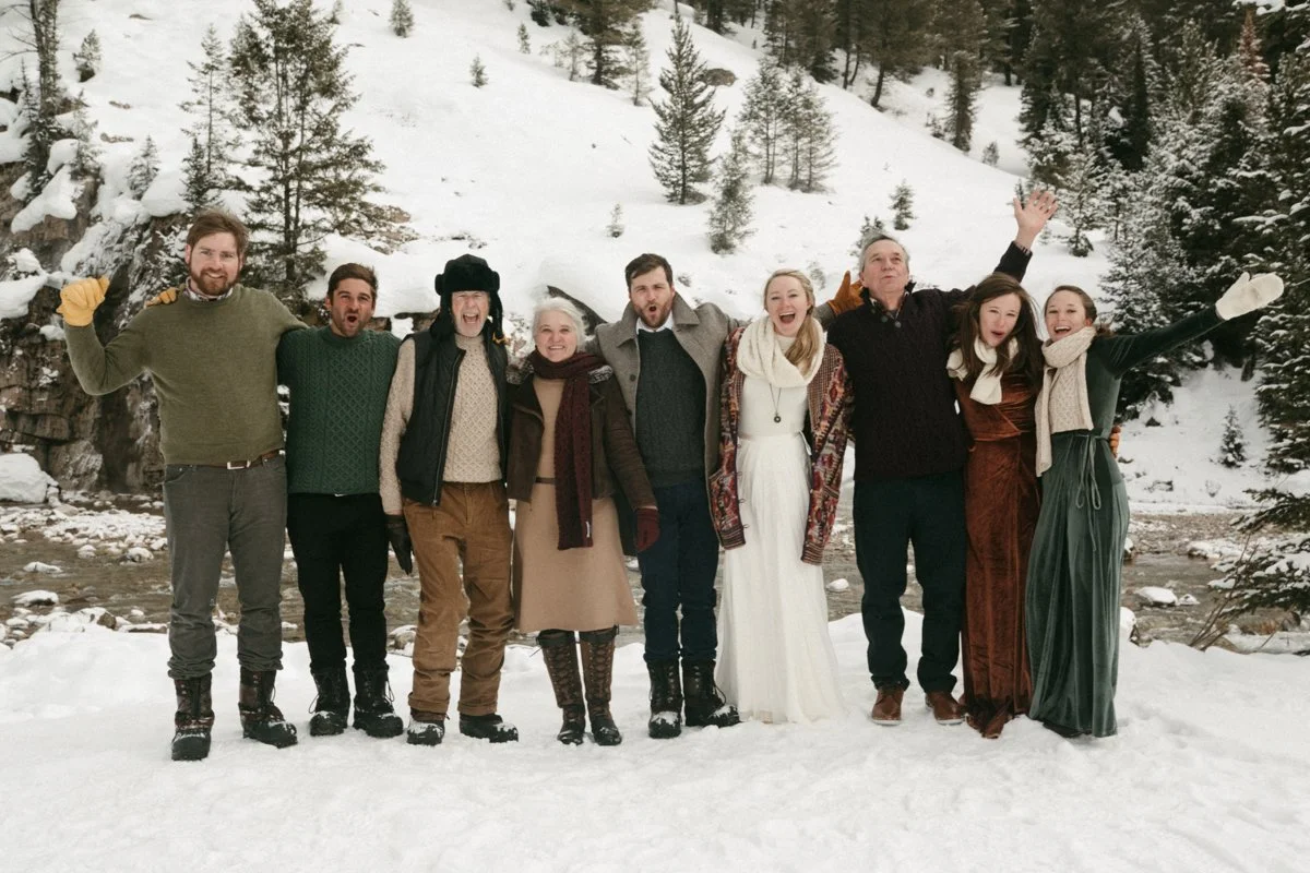 A wedding party poses for a photo next to a snowy river after a couple's Jackson Hole winter wedding ceremony